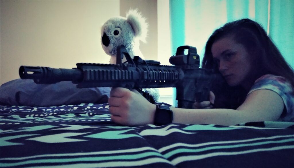 A young girl with an AR-15 rifle behind a bed